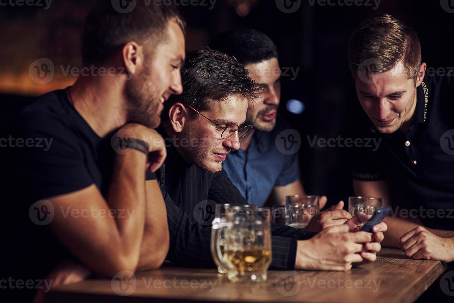 Funny content on smartphone. Three friends resting in the pub with beer in hands. Having conversation photo