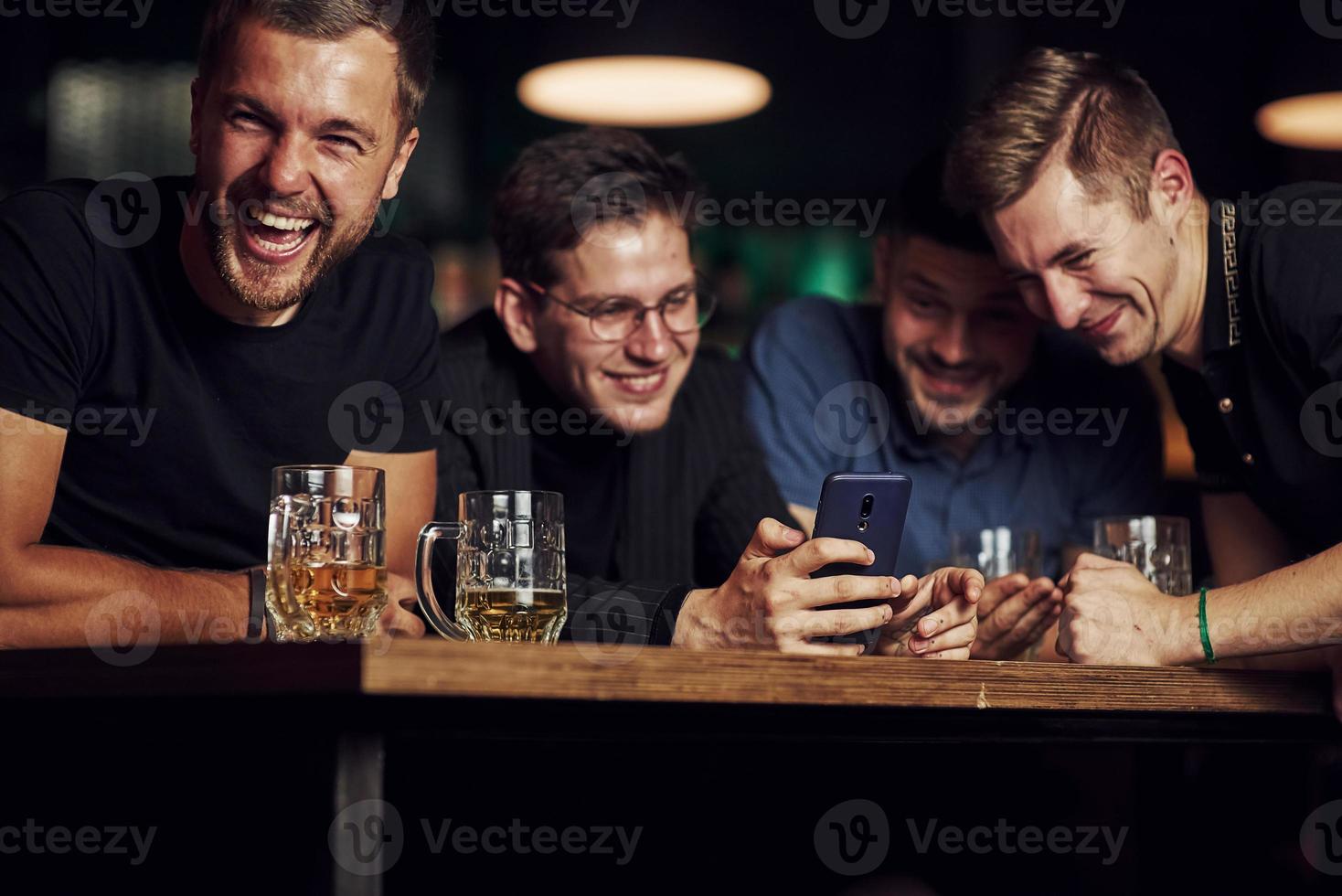 Funny content on smartphone. Three friends resting in the pub with beer in hands. Having conversation photo