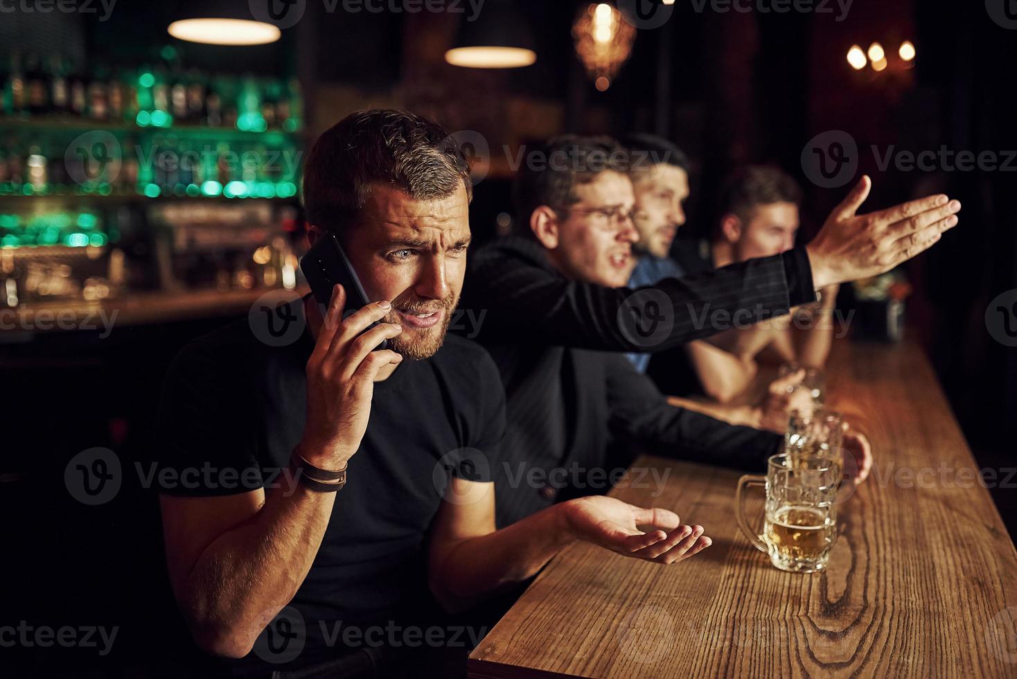 el hombre habla por teléfono. tres aficionados al deporte en un bar viendo fútbol. con cerveza en las manos foto