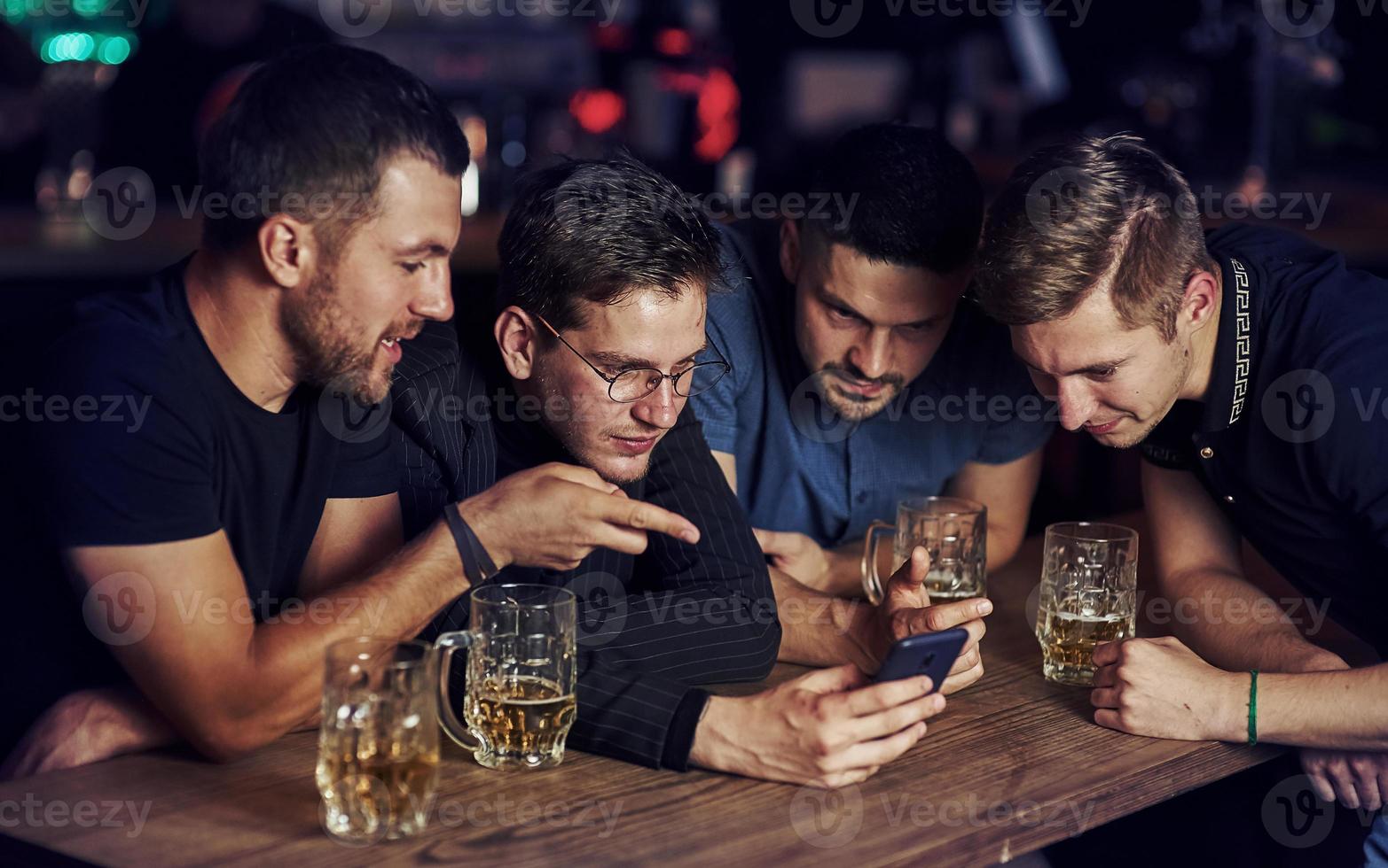 Funny content on smartphone. Three friends resting in the pub with beer in hands. Having conversation photo