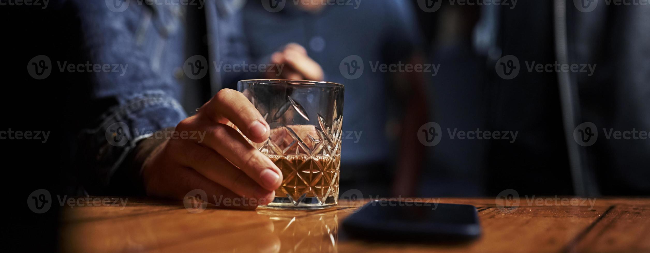 Close up view of glass with alcohold. Man's hand holds it. Smartphone on the table. Horizontal photo