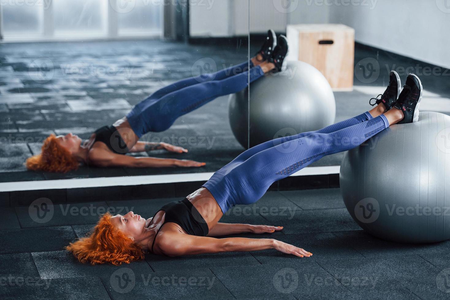 Using silver colored exercise ball. Sporty redhead girl have fitness day in gym at daytime. Muscular body type photo