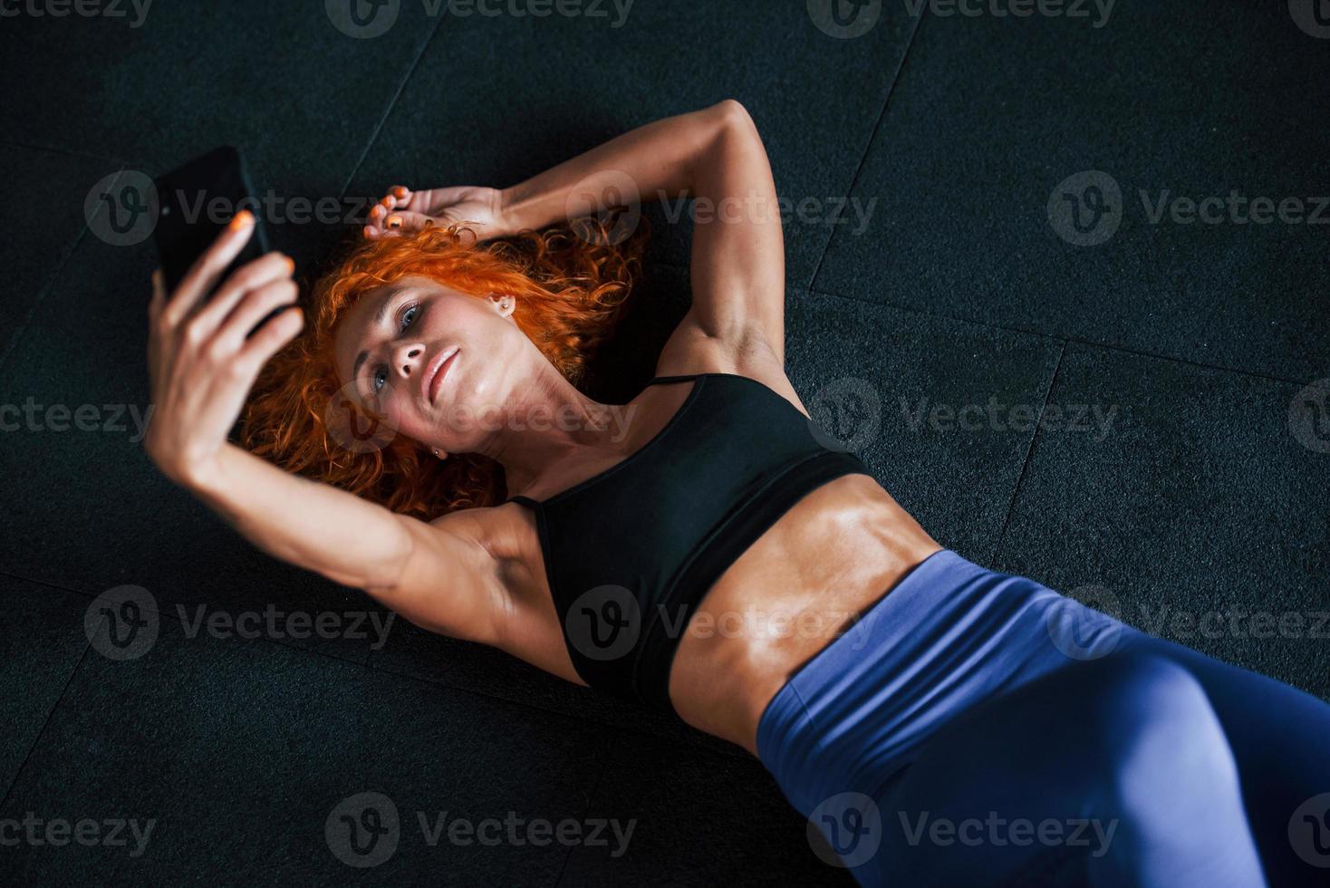 Taking a selfie with phone. Sporty redhead girl have fitness day in gym at daytime. Muscular body type photo