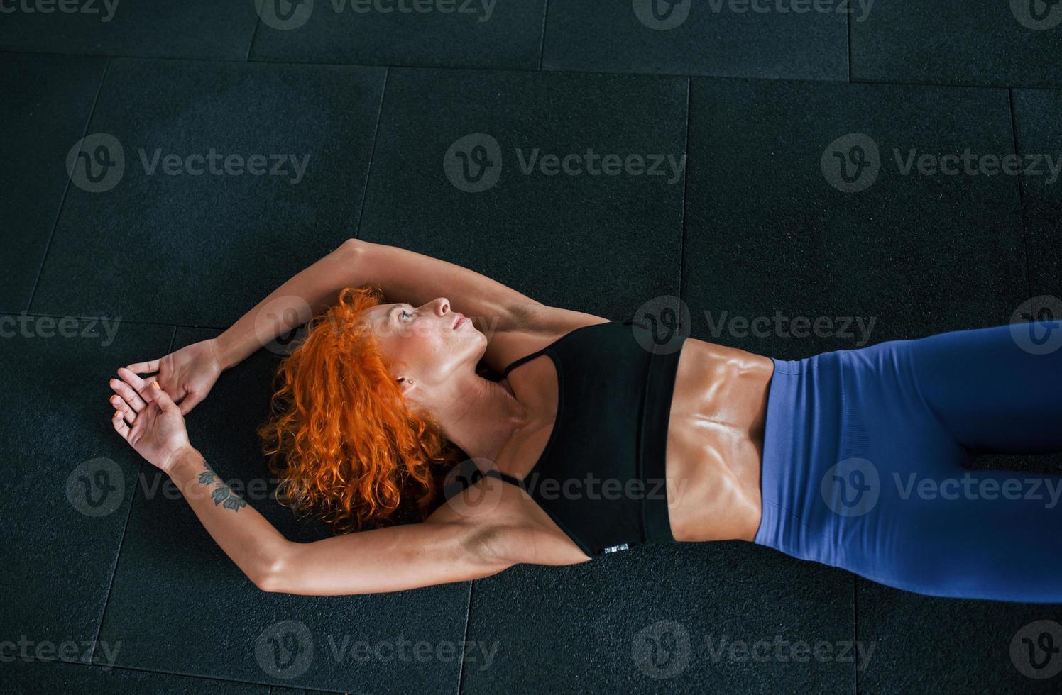 Lying down. Sporty redhead girl have fitness day in gym at daytime. Muscular body type photo