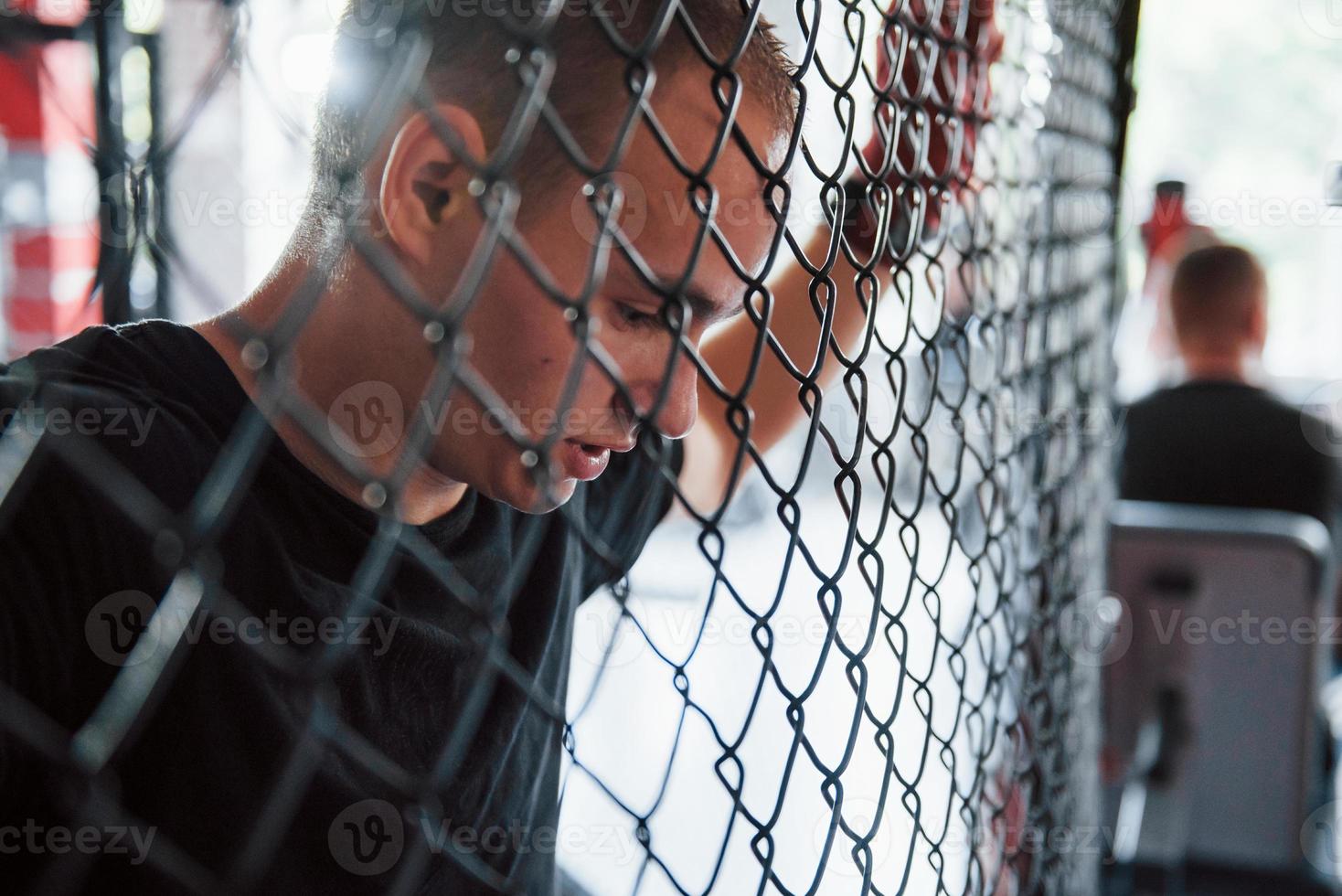 Side view. Taking a break. Sportsman at boxing ring have exercise. Leaning on the fence photo