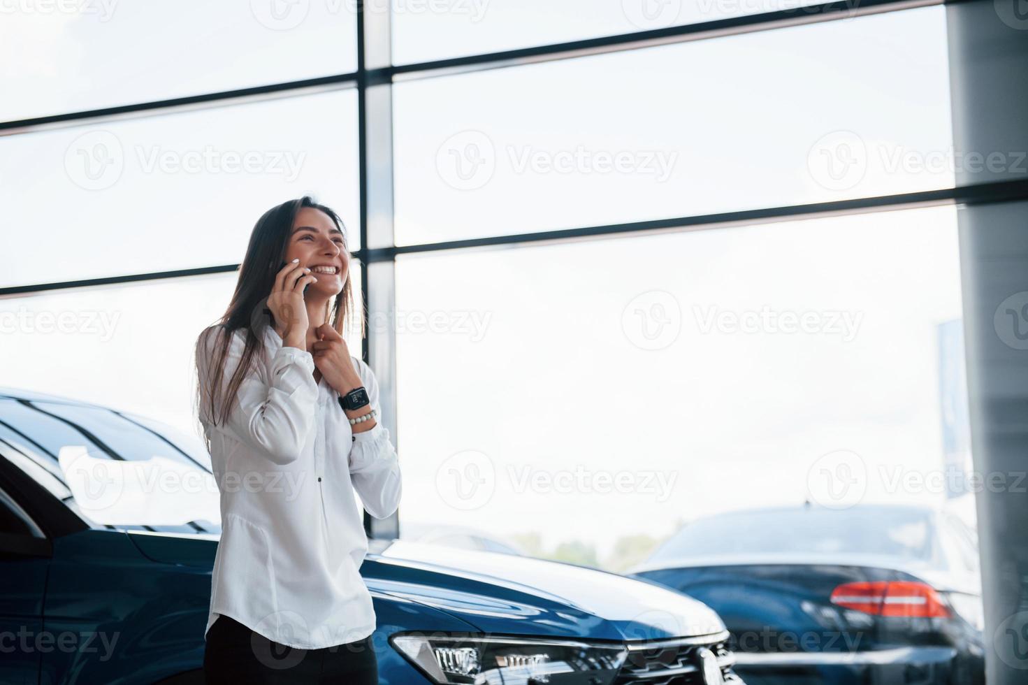 tener una conversación por teléfono. una joven con ropa oficial blanca se para frente a un automóvil azul en el interior foto