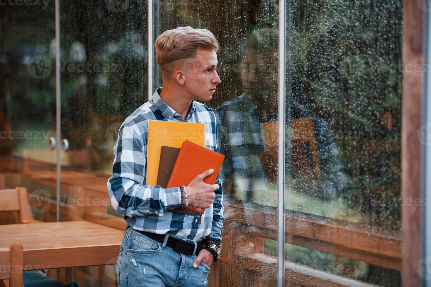 Holds notepads in hands. Portrait of young student that stands in cafe at rainy day. One person photo