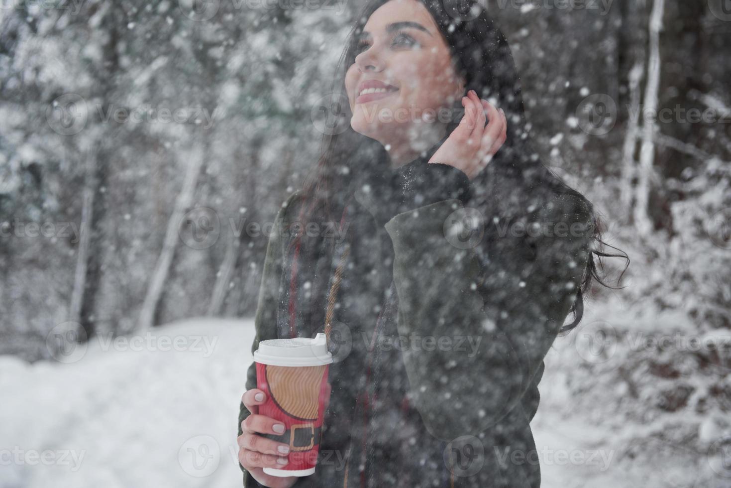 Snow is falling down from the sky. Girl in warm clothes with cup of coffee have a walk in the winter forest photo