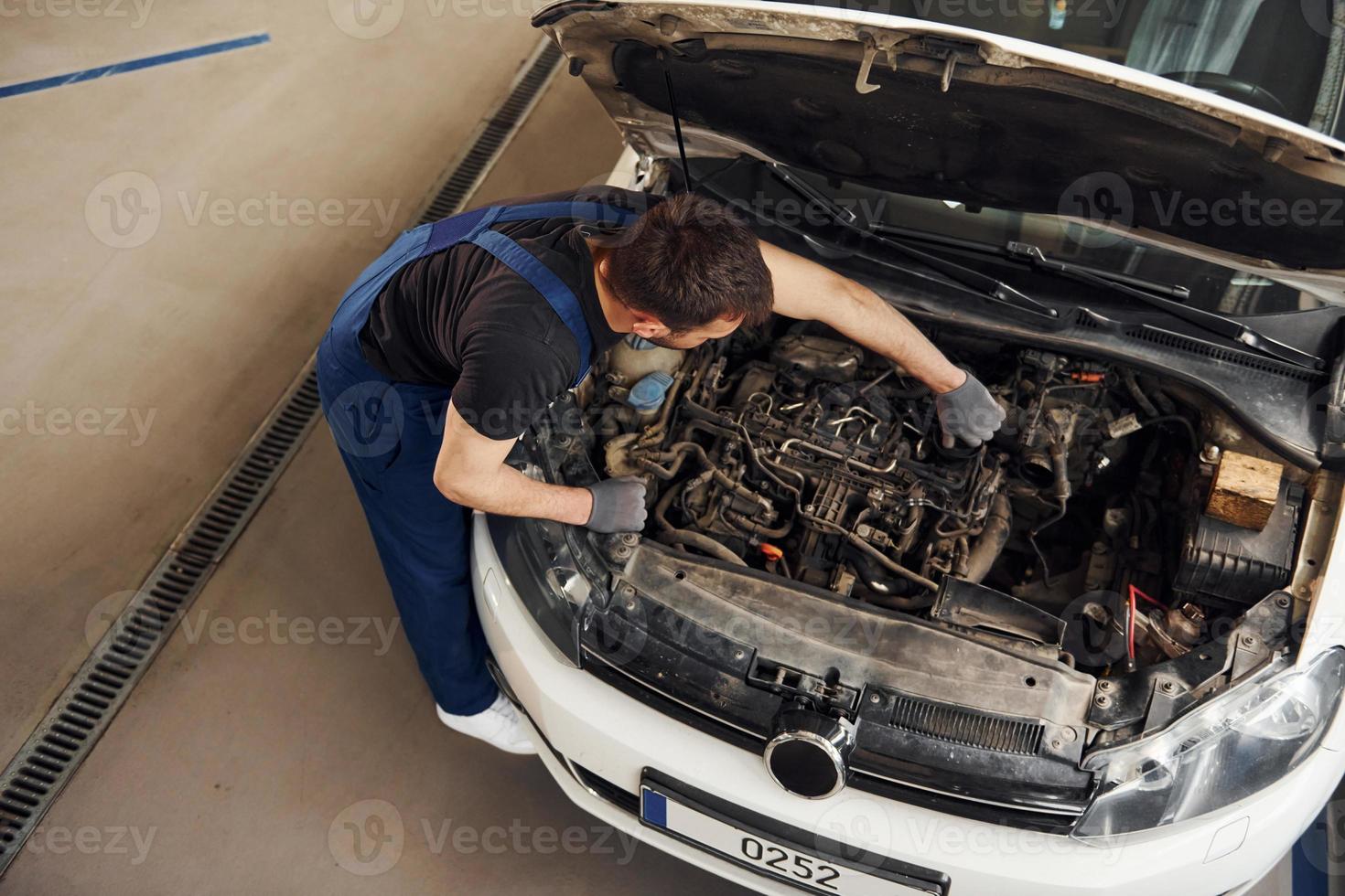 Under the hood. Man in work uniform repairs white automobile indoors. Conception of automobile service photo