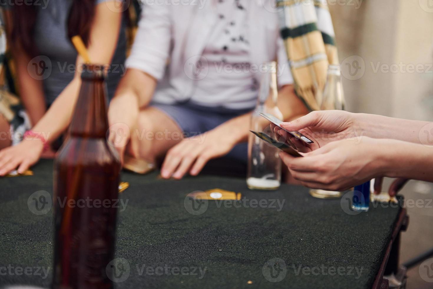 Sitting by table and playing card game. Group of young people in casual clothes have a party at rooftop together at daytime photo