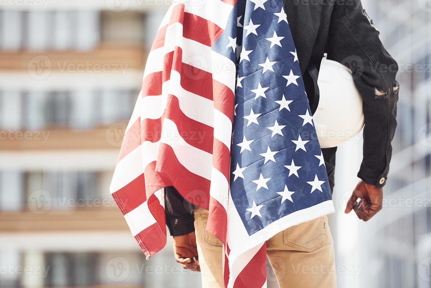 View from behind. Patriot holding USA flag. Conception of pride and freedom. Young african american man in black jacket outdoors in the city standing against modern business building photo