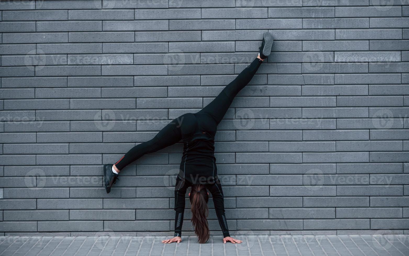 Young sportive girl in black sportswear doing hard handstand exercises outdoors near gray wall photo