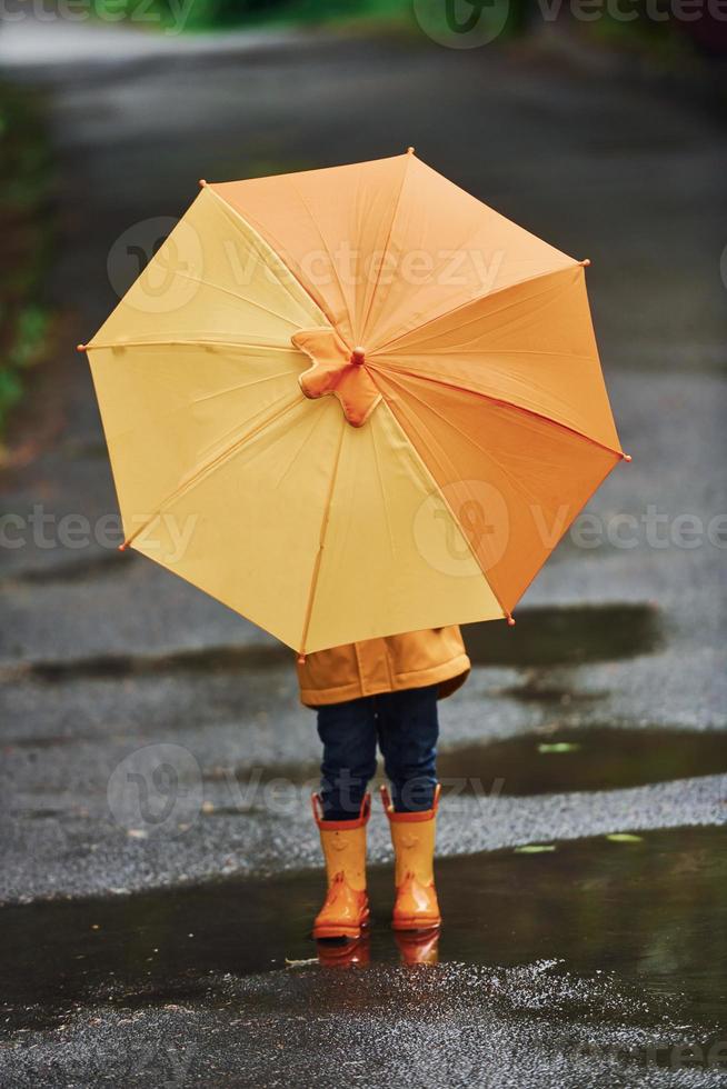 Kid in yellow waterproof cloak, boots and with umbrella playing outdoors after the rain photo