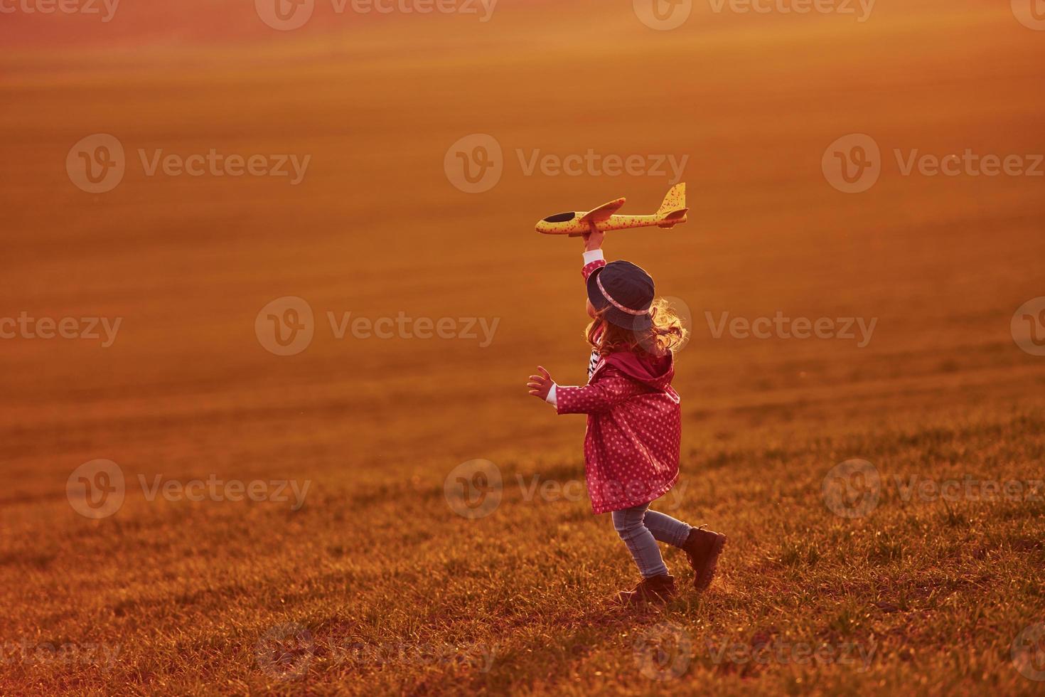 Illuminated by orange colored sunlight. Cute little girl have fun with toy plane on the beautiful field at daytime photo