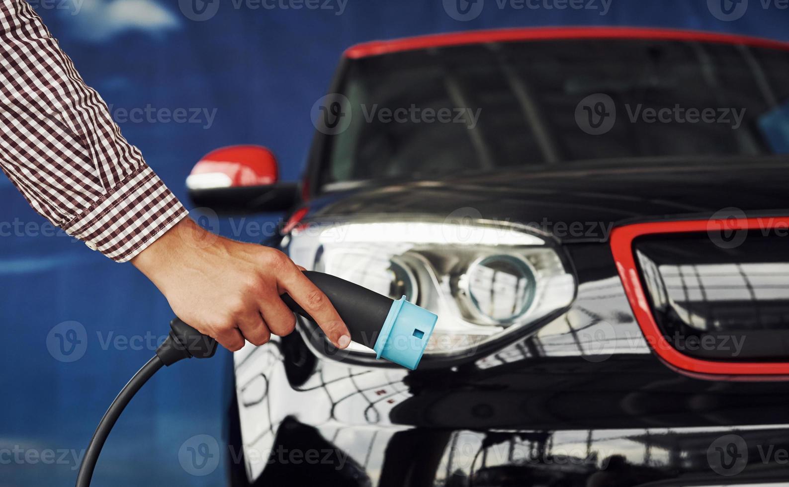 Man stands against an electric car and holds the charging cable photo