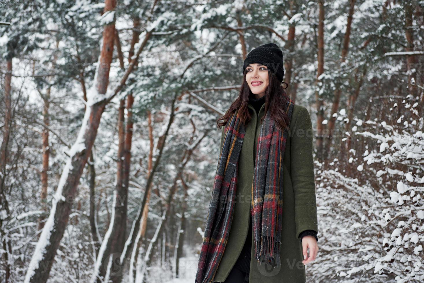 Standing in the middle of the woods. Cheerful young girl in warm clothes have a walk in the winter forest at daytime photo