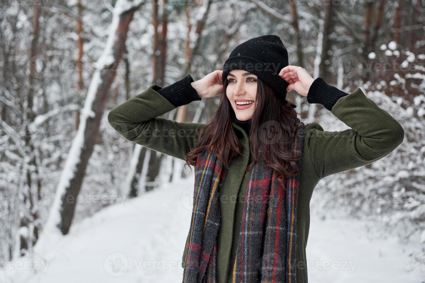 Touches the black hat. Cheerful young girl in warm clothes have a walk in the winter forest at daytime photo