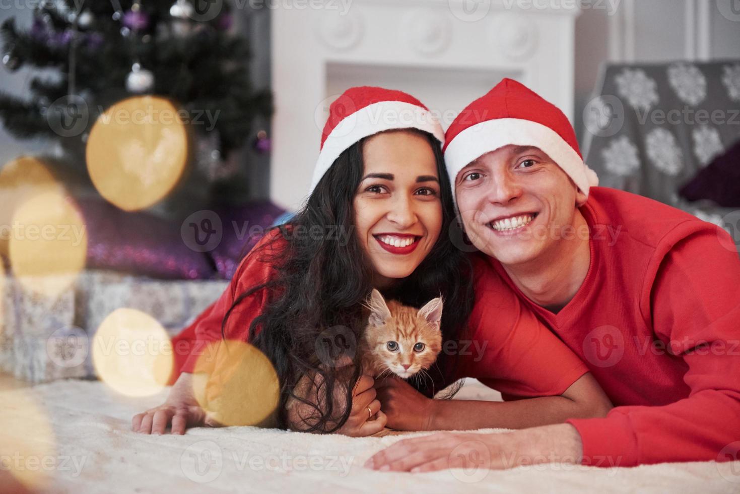 Lying on the floor. Everyone including cat looking into the camera. Portrait of couple with little kitty celebrates holidays in new year clothes photo