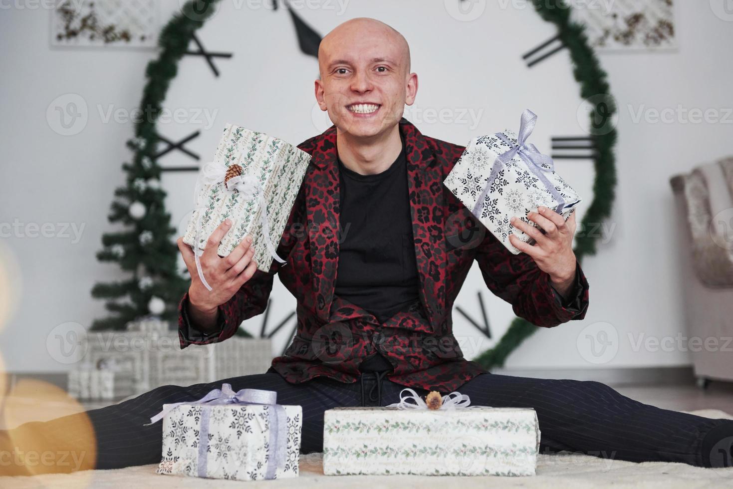 Holding two presents in hands. Man with big clock behind him sitting with New year gift boxes in holiday clothings photo