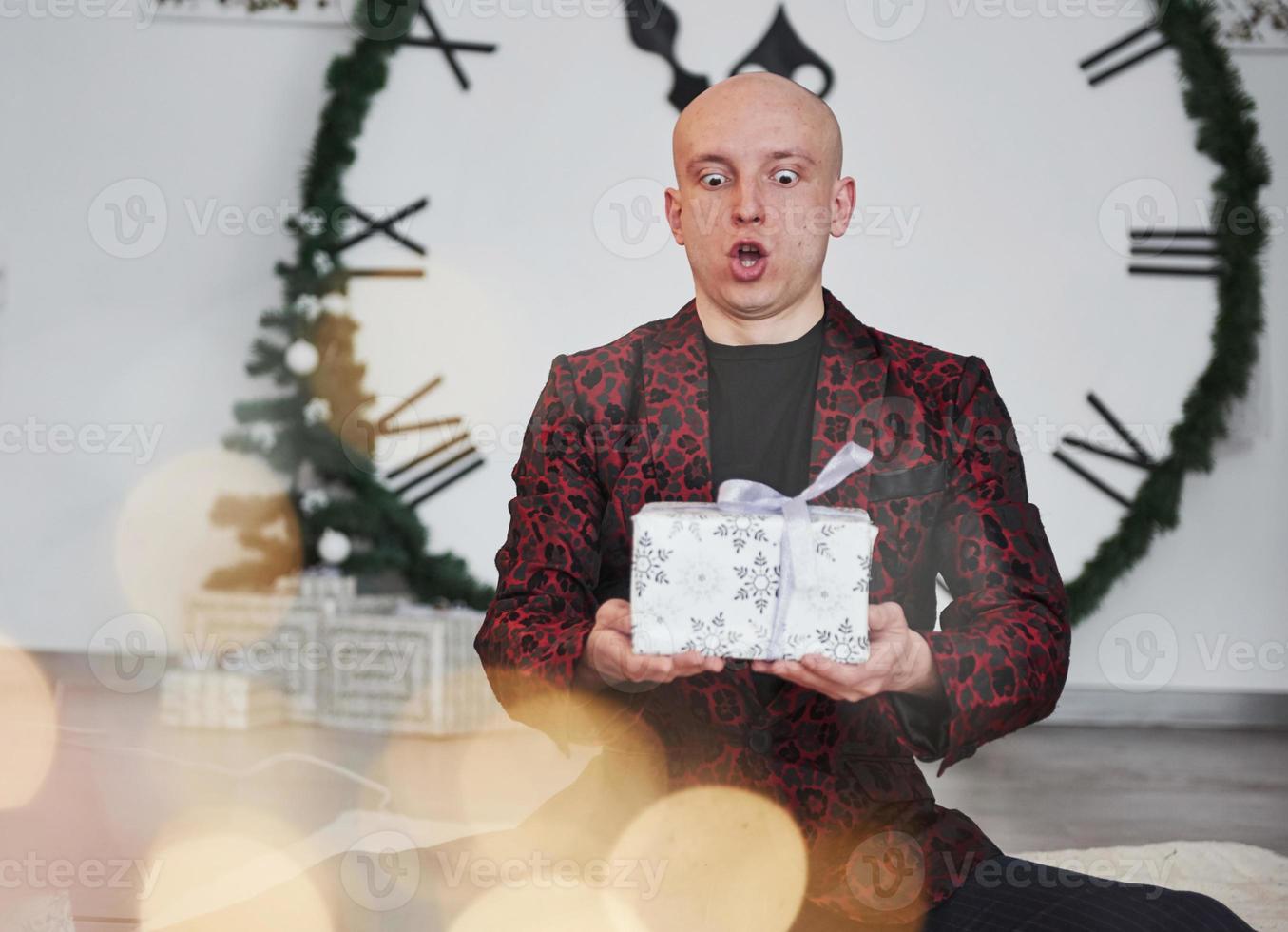 Face expression. Man with big clock behind him sitting with New year gift boxes in holiday clothings photo