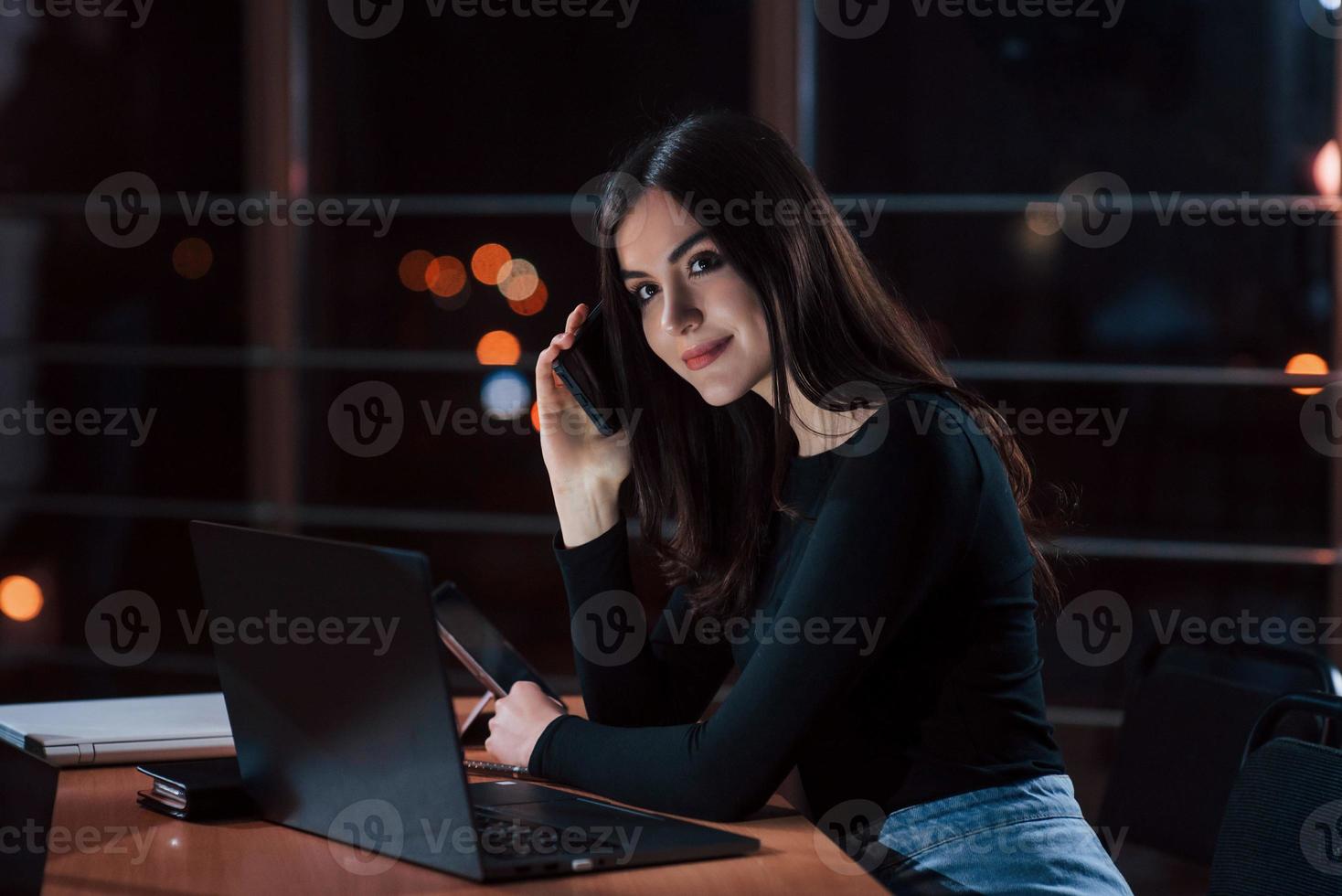 gran teléfono inteligente cerca de la oreja. atractiva mujer de negocios morena trabaja sola en la oficina por la noche foto