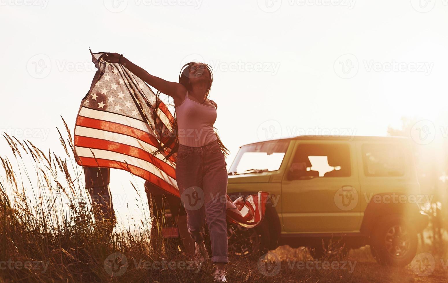 la chica corre hacia adelante. los amigos tienen un buen fin de semana al aire libre cerca de su auto verde con bandera de estados unidos foto