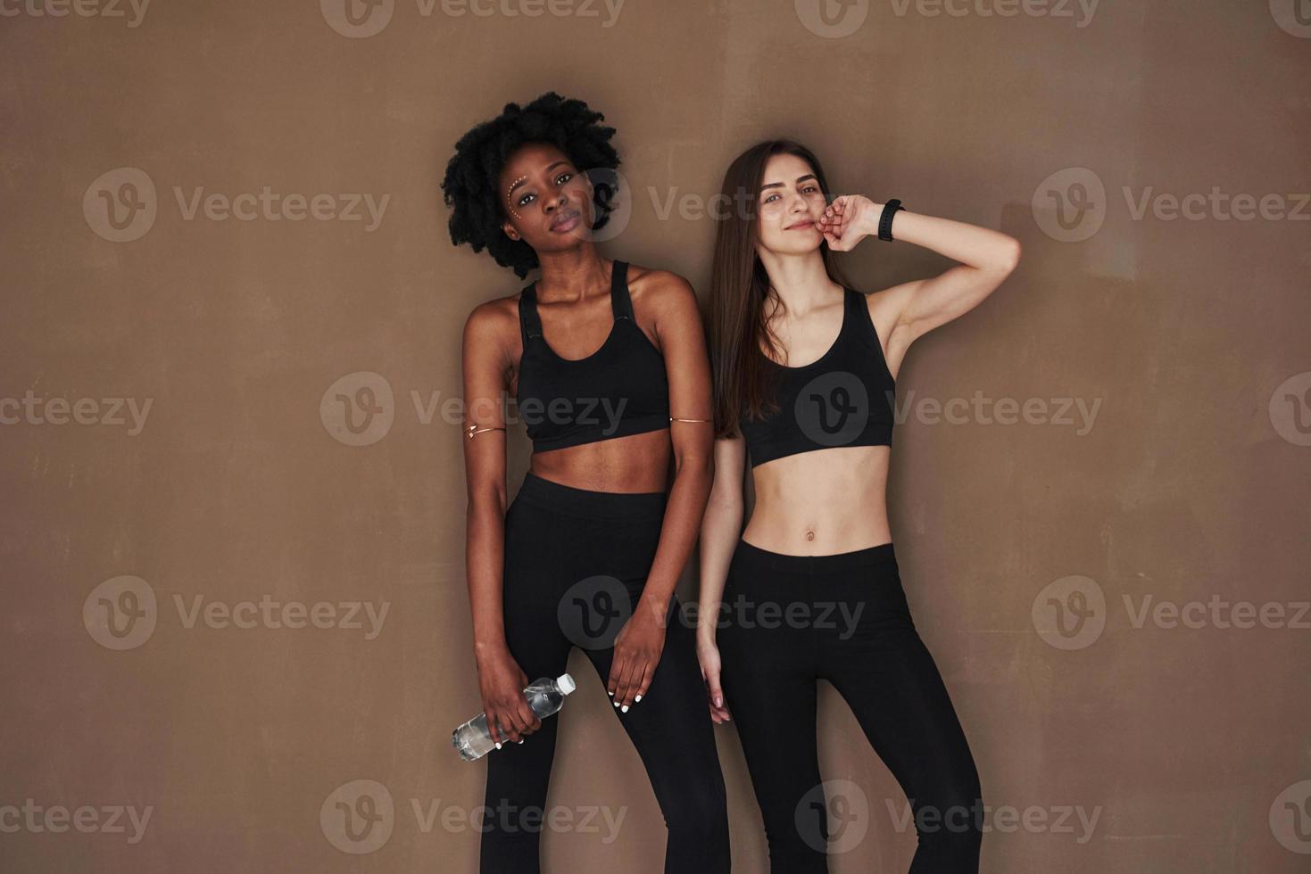 Young and active. Two multi ethnic female friends stands in the studio with brown background photo