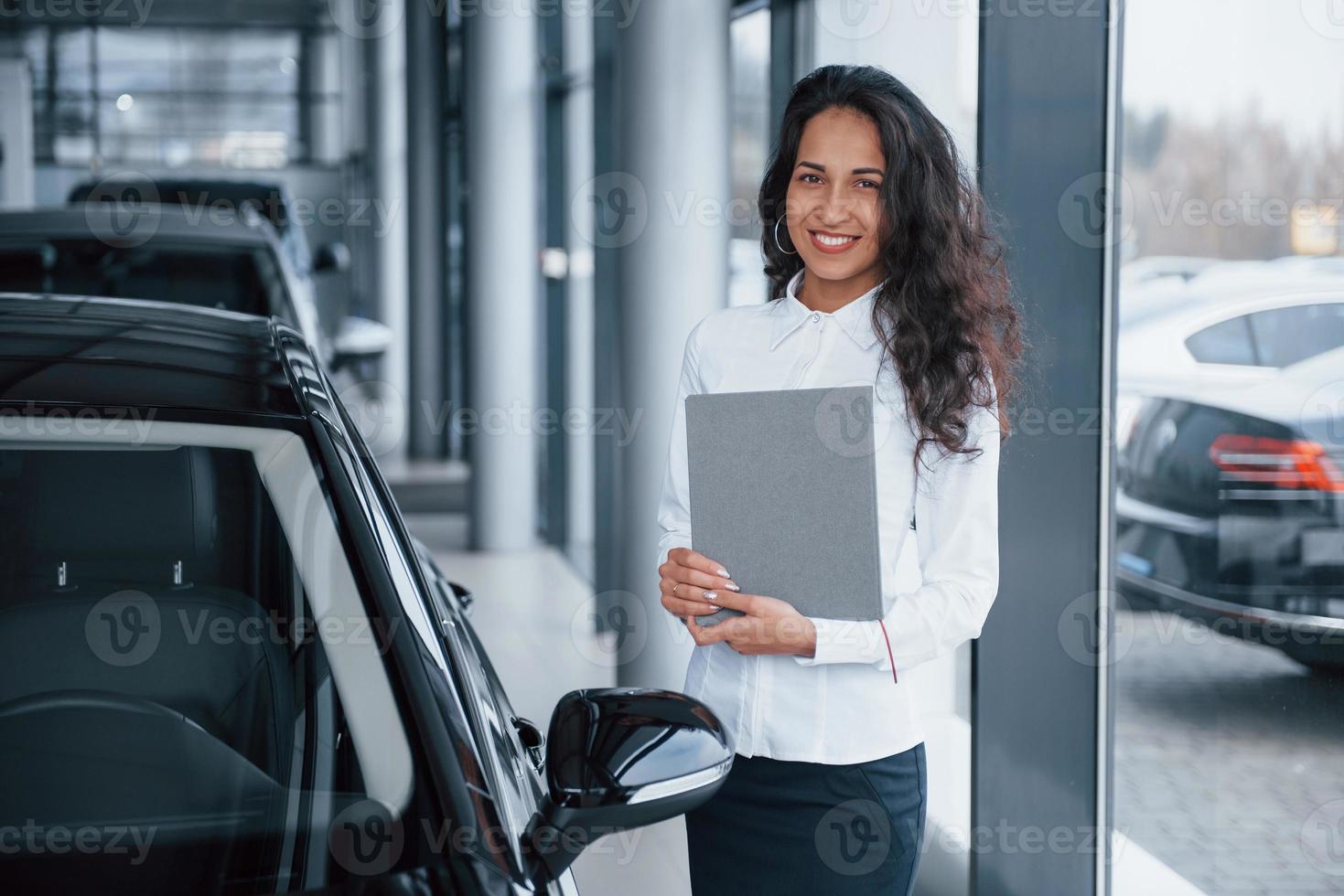 How can I help you. Curly haired female manager stands near the car in automobile salon photo