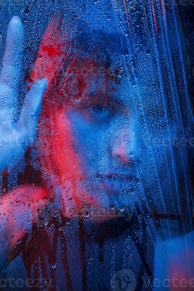 Red and blue lights on woman. Studio shot in dark studio with neon light. Portrait of beautiful girl behind wet glass photo