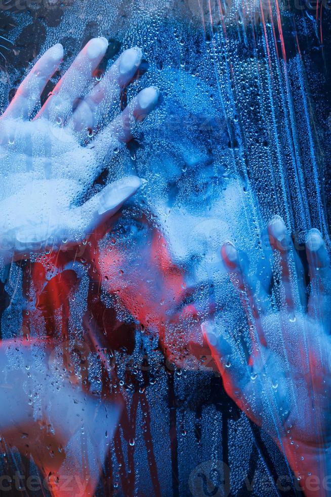 Looking with interest. Studio shot in dark studio with neon light. Portrait of beautiful girl behind wet glass photo