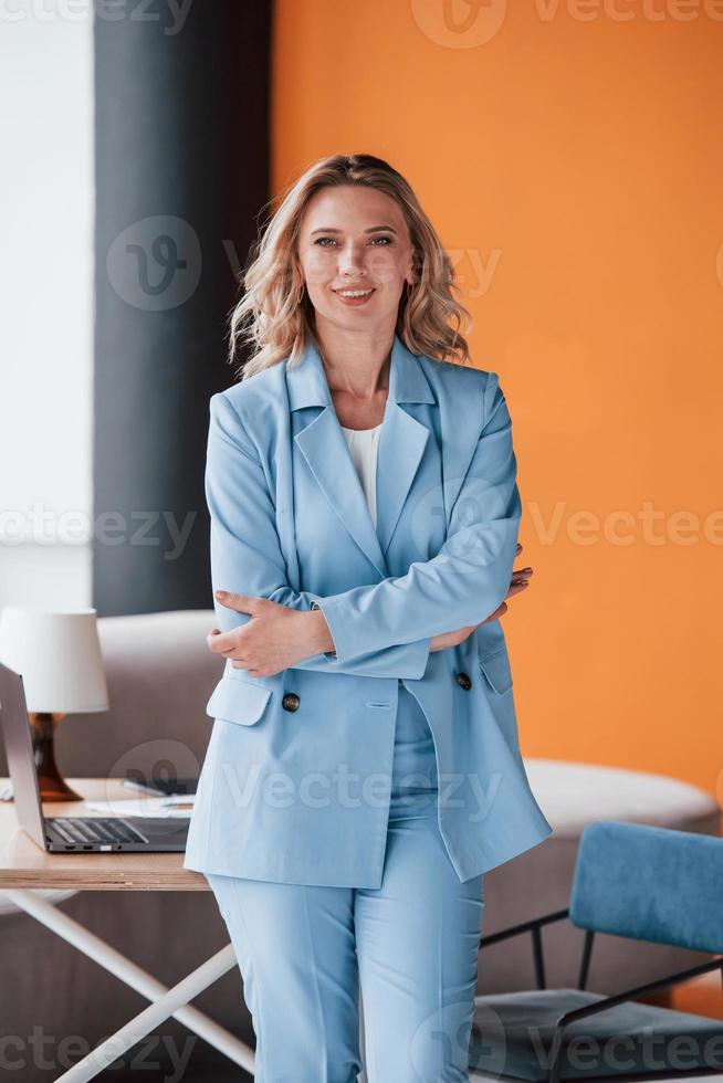 We have open vacancy, welcome. Businesswoman with curly blonde hair indoors in room with orange colored wall and wooden table photo