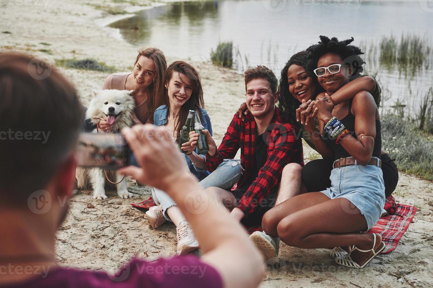 Everyone looks in the camera. Group of people have picnic on the beach. Friends have fun at weekend time photo