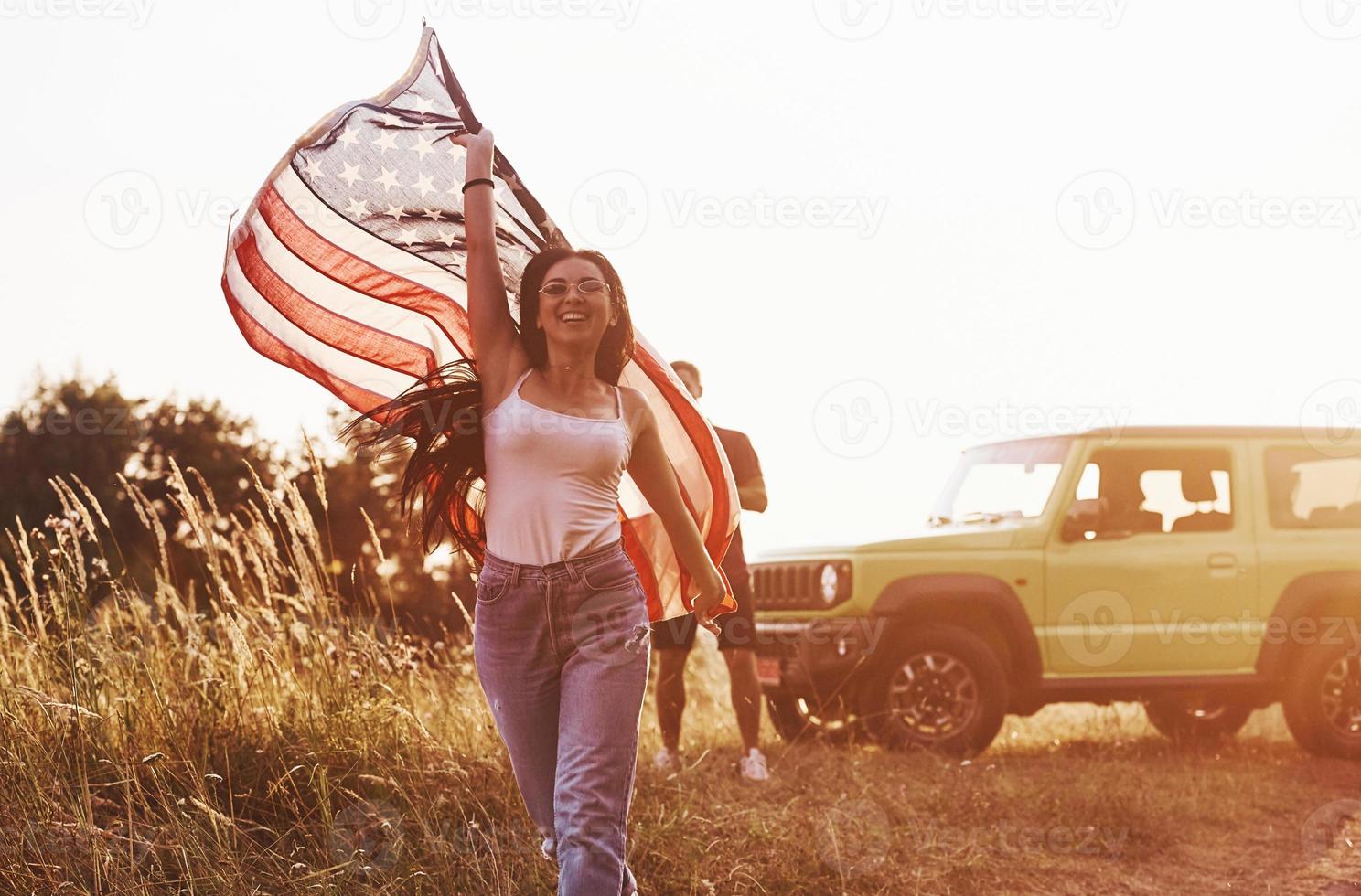 Girl runs forward. Friends have nice weekend outdoors near theirs green car with USA flag photo