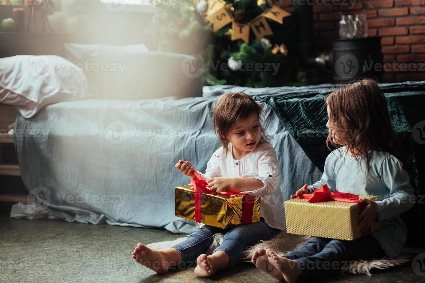 I'll open my box first. Christmas holidays with gifts for these two kids that sitting indoors in the nice room near the bed photo