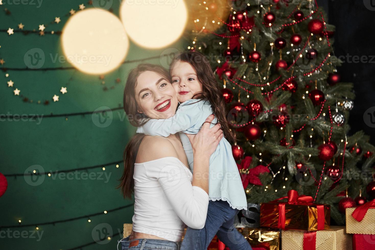 Perfect lighting. Cheerful mother and daughter hugging each other near the Christmas tree that behind. Cute portrait photo