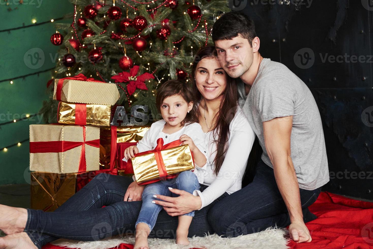 Lovely family sits near the Christmas tree with gift boxes on winter evening, enjoying the time spending together photo