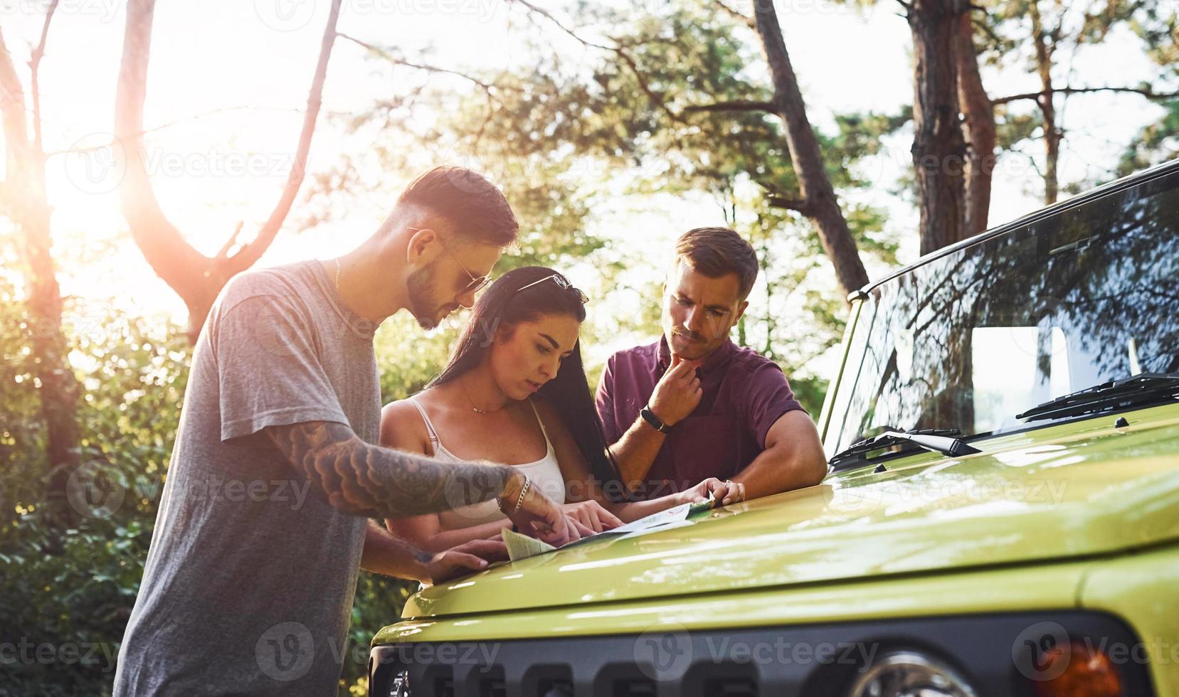 Young friends reading map that is on the hood of modern green jeep in the forest photo