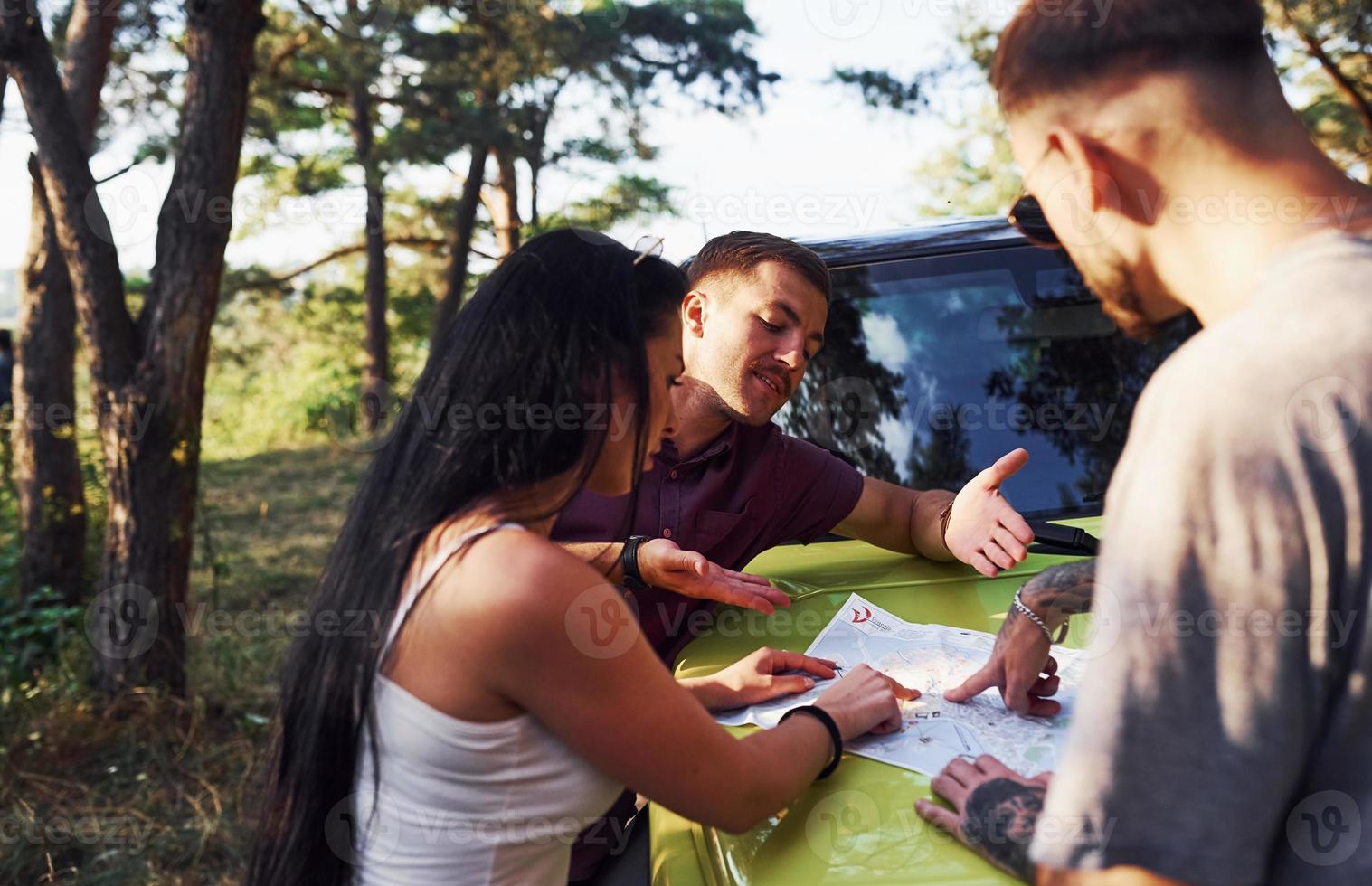 Young friends reading map that is on the hood of modern green jeep in the forest photo