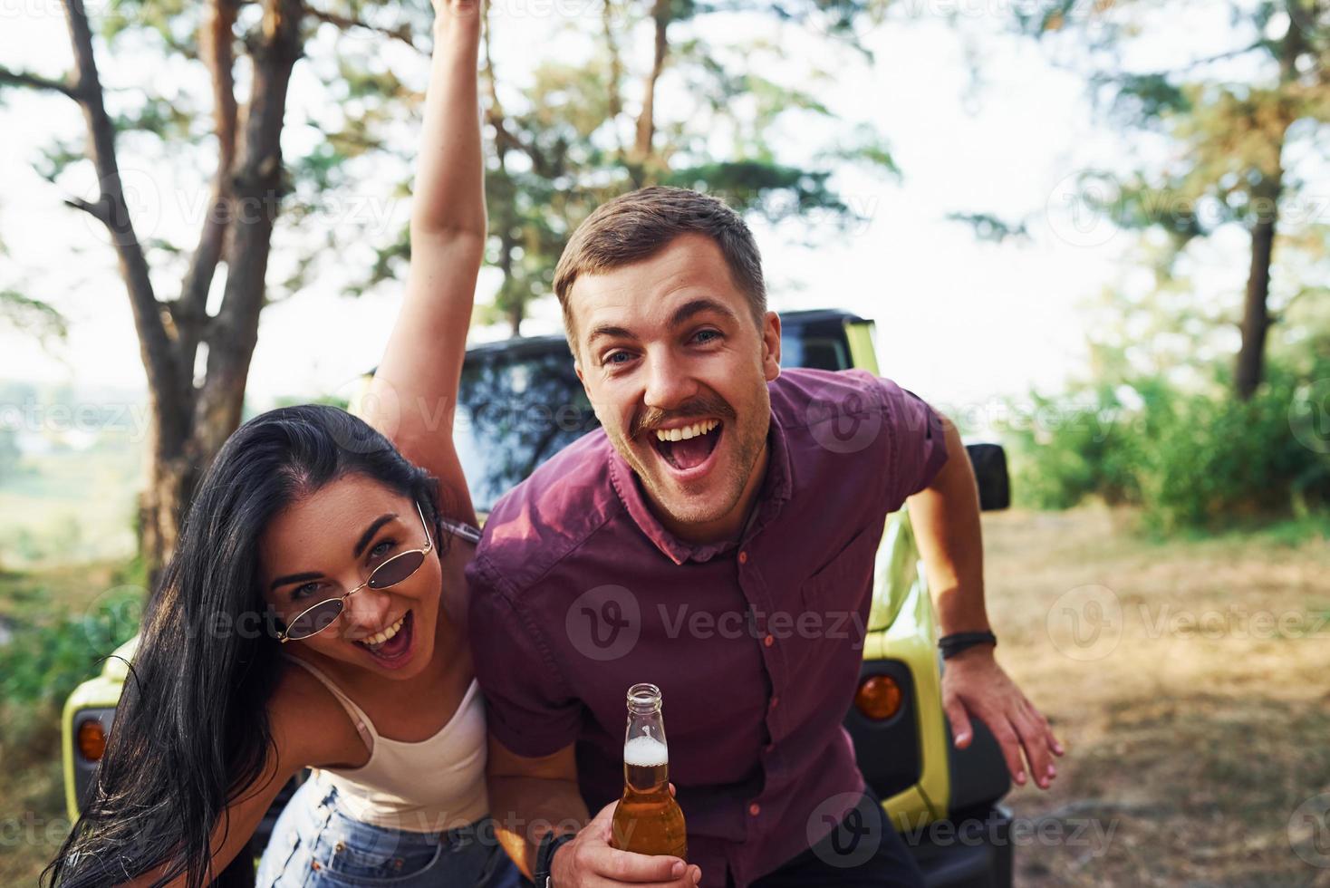 sonriendo y divirtiéndose. un par de jóvenes con alcohol se divierten en el bosque. jeep verde detrás foto