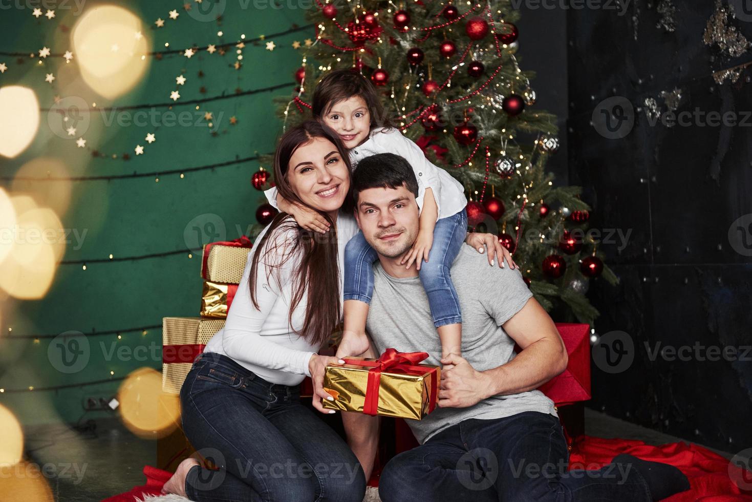 Posing for the picture. Lovely family sits near the Christmas tree with gift boxes on winter evening, enjoying the time spending together photo