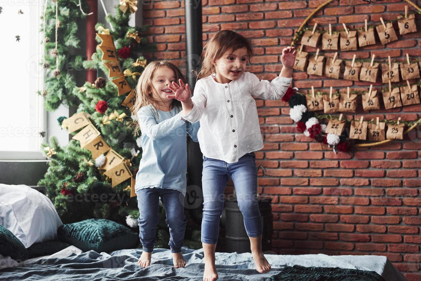 Letters on the tree means merry Christmas. Cheerful kids having fun and jumping on the bed with decorative holiday background photo