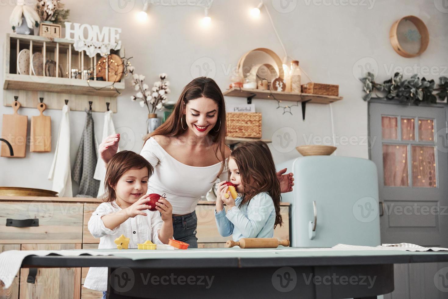Eating fresh fruits. Young beautiful woman feeds two kids with apples while they sitting near the table with toys photo