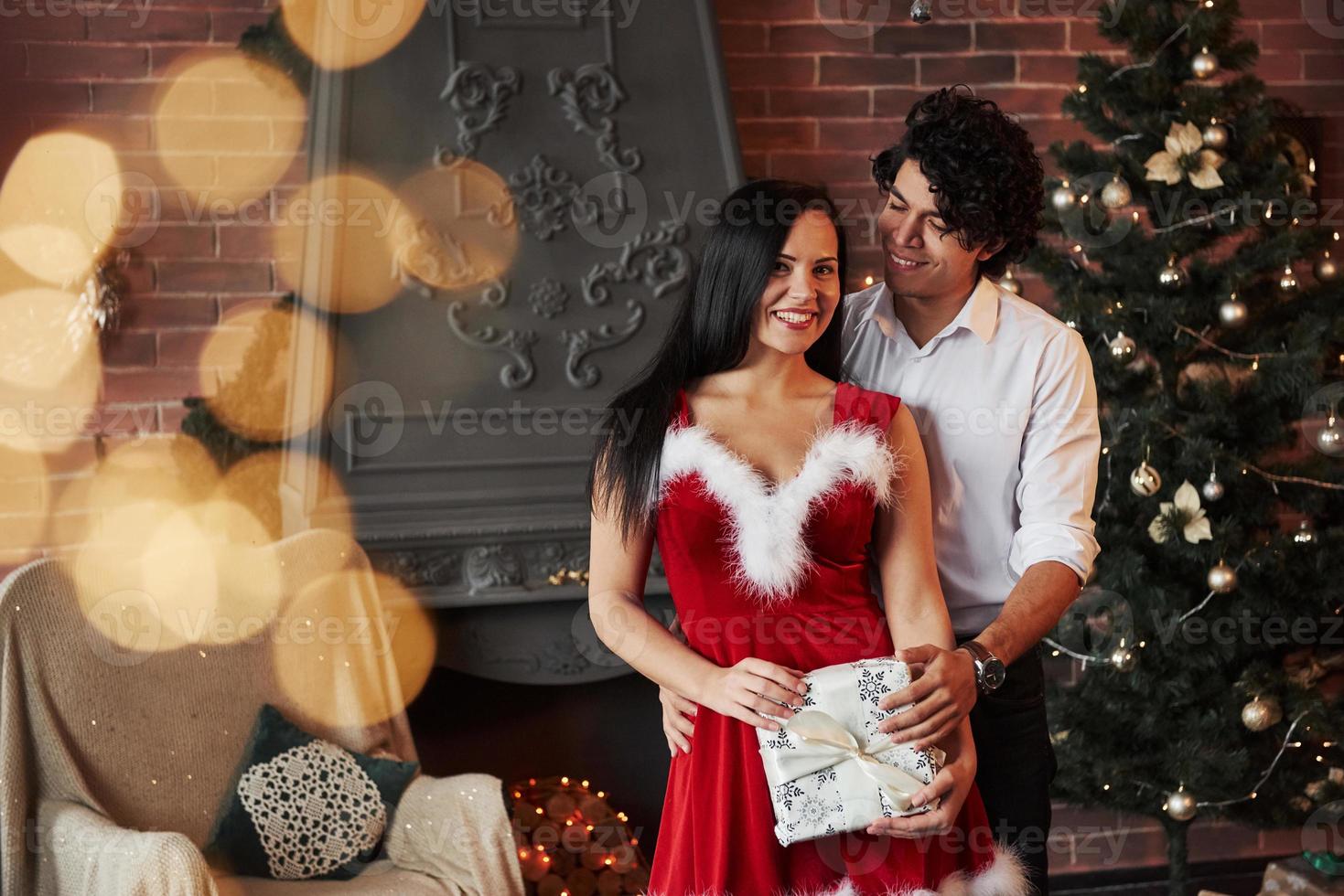Holding the holiday gift in hands. Beautiful couple celebrating New year in the decorated room with Christmas tree and fireplace behind photo