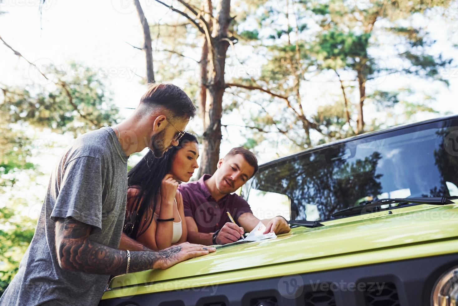 Young friends reading map that is on the hood of modern green jeep in the forest photo