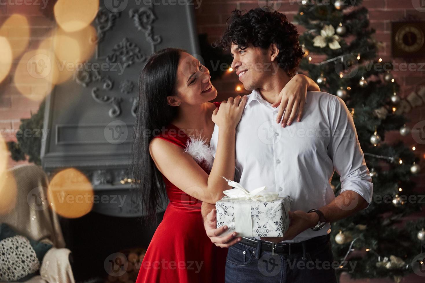 Such a lovely pair. Beautiful couple celebrating New year in the decorated room with Christmas tree and fireplace behind photo