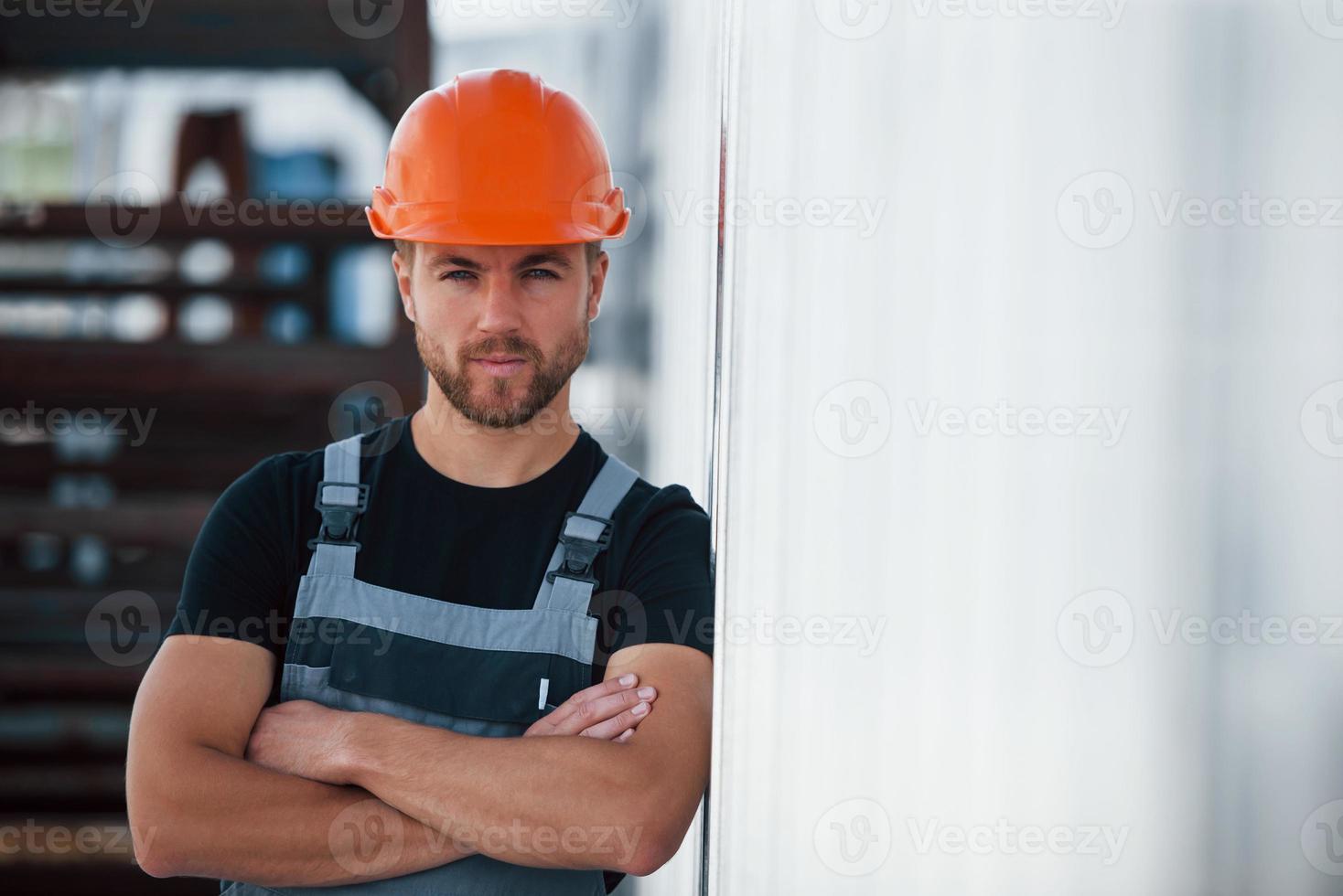 Leaning on the wall. Serious industrial worker indoors in factory. Young technician with orange hard hat photo