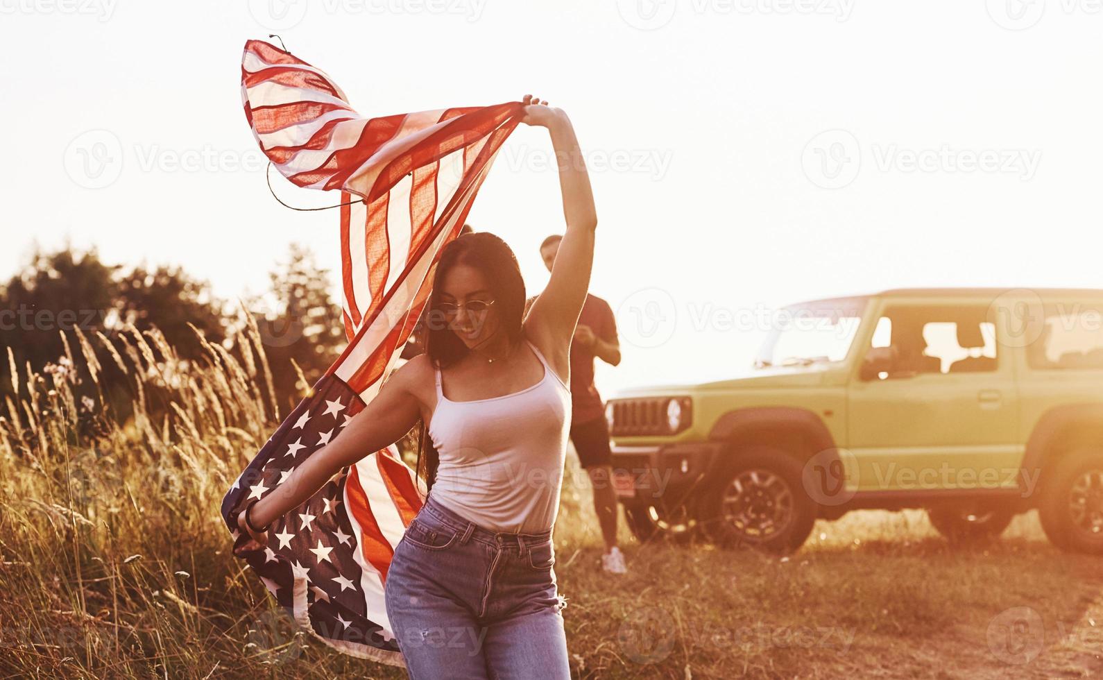 Girl runs forward. Friends have nice weekend outdoors near theirs green car with USA flag photo