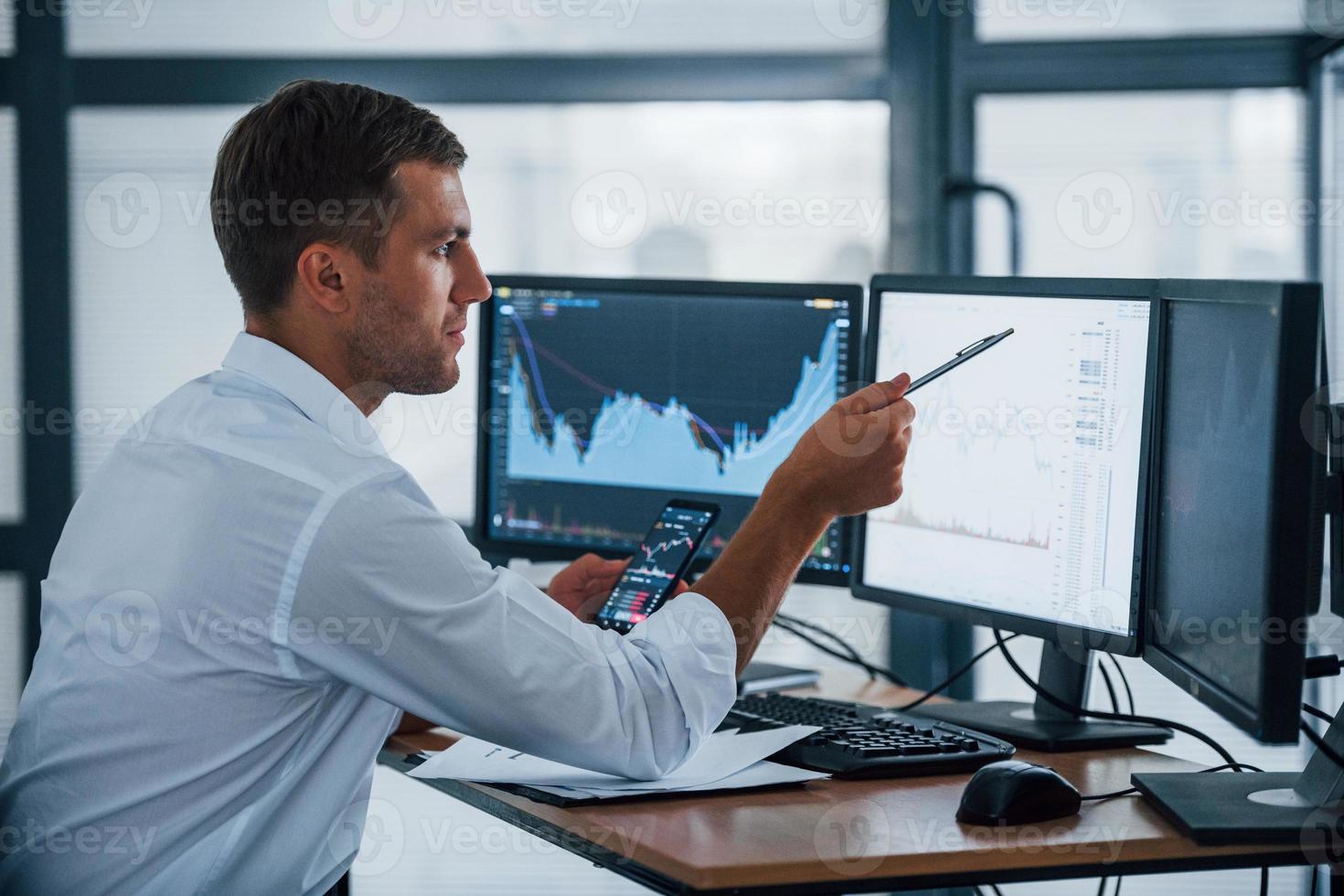 Young businessman in formal clothes is in office with multiple screens. Conception of exchange and money photo