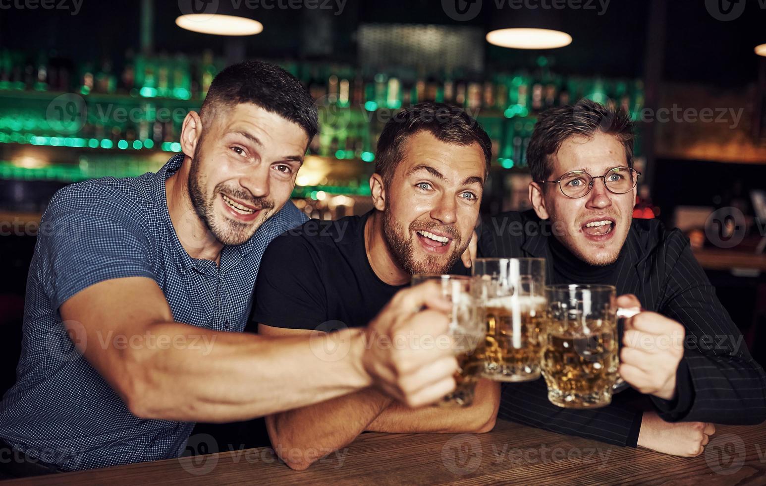 golpeando vasos. tres aficionados al deporte en un bar viendo fútbol. con cerveza en las manos foto