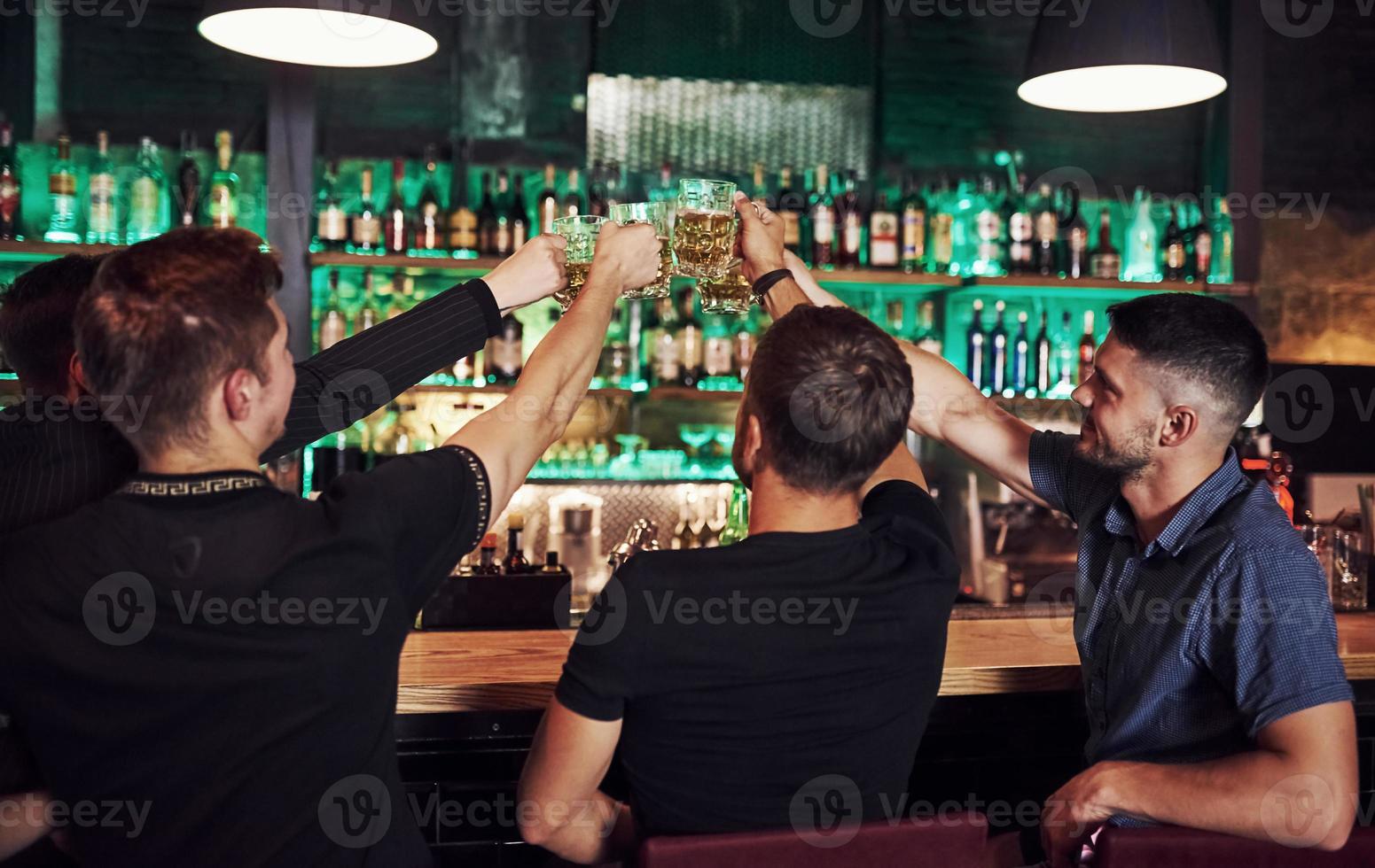 Many of bottles with alcohol on the shelves. Friends resting in the pub with beer in hands. Having conversation photo